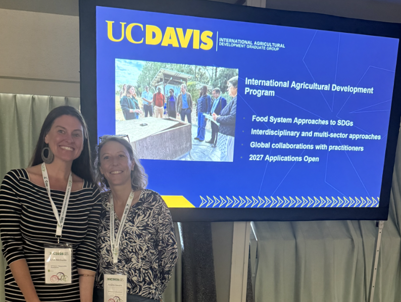 Two smiling conference attendees wearing badges before a UC Davis presentation screen (photo)