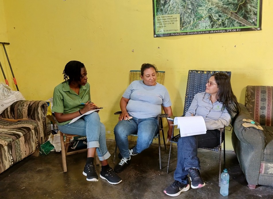 Three people seated in a small yellow-walled room in conversation, holding notebooks and papers