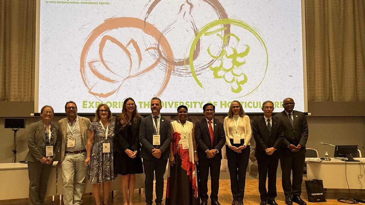 Eleven people in formal attire stand on a conference stage before a large projected logo